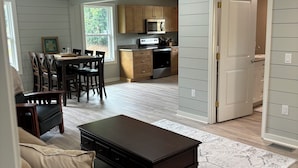 A spacious living area with a sofa, a coffee table, and a dining table with chairs. A kitchen is visible in the background with wooden cabinets and a microwave.