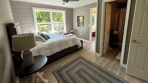 A bedroom with a bed, a nightstand, a lamp, a ceiling fan, and a window with a view of greenery.