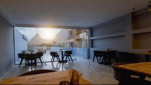 A modern interior with a large glass window offering a view of pyramids. There are tables and chairs arranged for dining. - Habu Pyramids View Hotel (Giza)