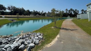 A paved pathway leading to a calm pond, surrounded by a grassy area and a row of trees.