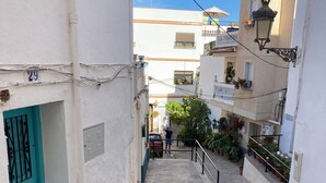 A narrow, paved street with white buildings, a blue door, and a staircase leading to a higher level.
