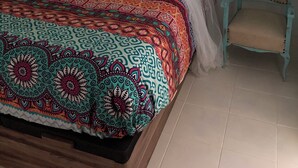 A bed with a colorful patterned bedspread and a canopy, a chair, and a teal wardrobe in a room with white walls.