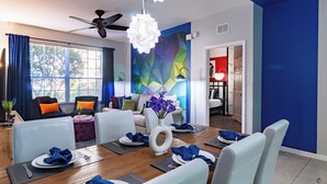 A modern dining area with a wooden table set for four, white chairs, and a ceiling fan. - Walt’s Vision – Disney-Inspired 3BR Condo (Kissimmee)