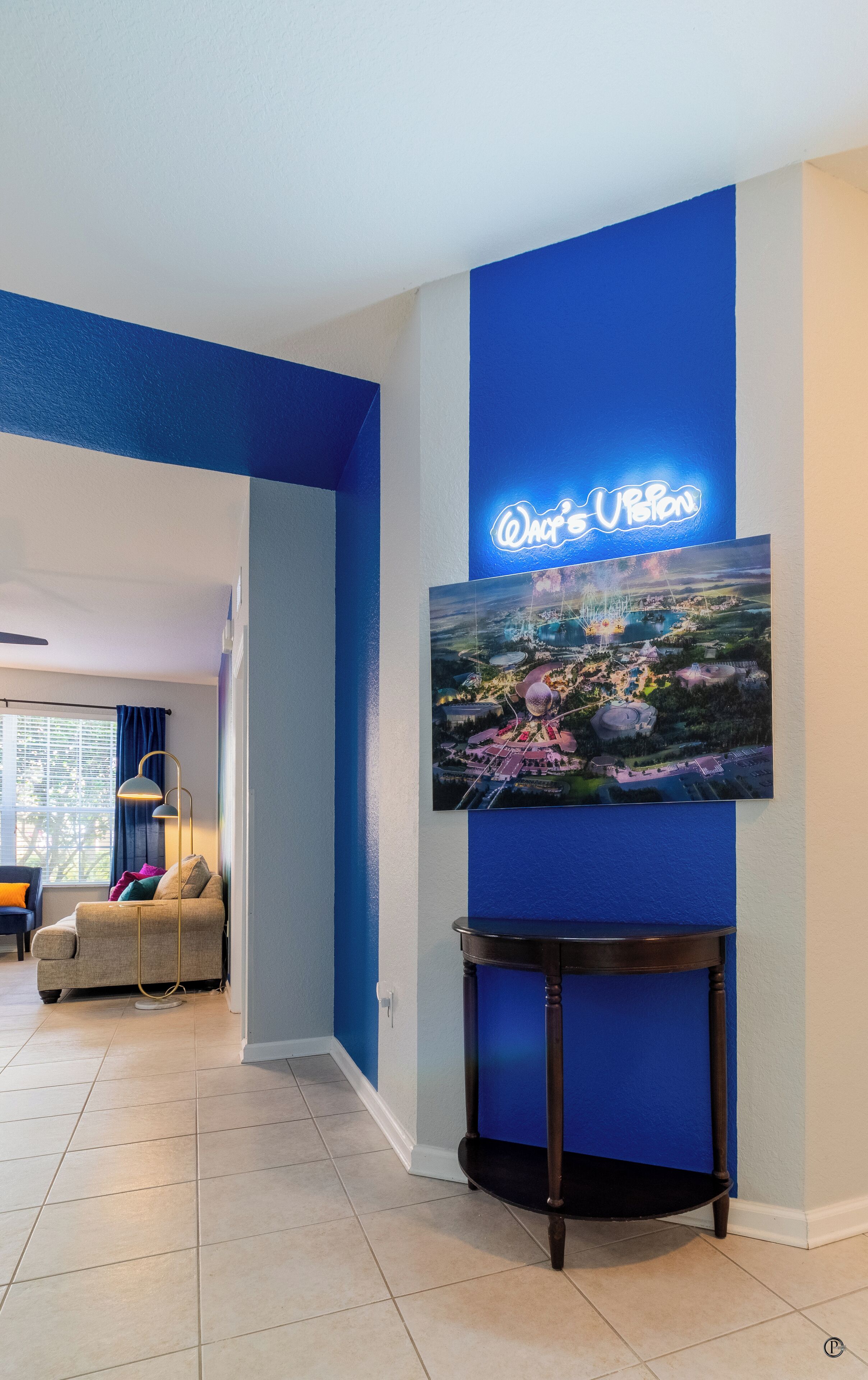 Una sala de estar moderna con un televisor grande montado en una pared azul, una zona de estar acogedora con un sofá y una lámpara, y piso de baldosas.