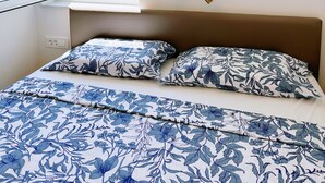 A bedroom with a bed, floral-patterned bedding, a window with a view of buildings and greenery, and a wall-mounted light fixture.