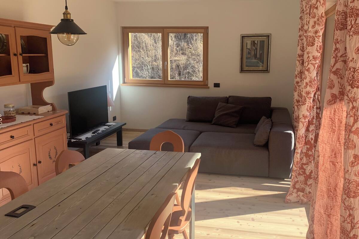 Un salon chaleureux avec une table à manger en bois, un canapé, un téléviseur et une fenêtre donnant sur des arbres.