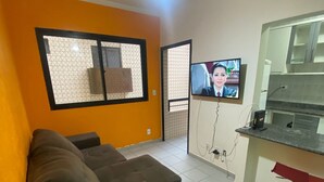 A living room with a sofa, a TV, a ceiling fan, and a kitchen area with a countertop and sink. - Foot on the sand beach of Guilhermina III (Praia Grande)