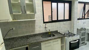 A kitchen with white cabinets, a granite countertop, a sink, a stove, and a window with blinds. - Foot on the sand beach of Guilhermina III (Praia Grande)