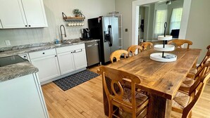 A modern kitchen with white cabinets, a stainless steel refrigerator, a coffee maker, and a dining table with wooden chairs. - Burnside Boathouse – Cozy 5 Bedroom Lake Retreat Steps from Marina & Local Eats (Burnside)