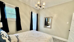 A bedroom with a bed, a chandelier, a mirror, and a window with curtains. - Burnside Boathouse – Cozy 5 Bedroom Lake Retreat Steps from Marina & Local Eats (Burnside)