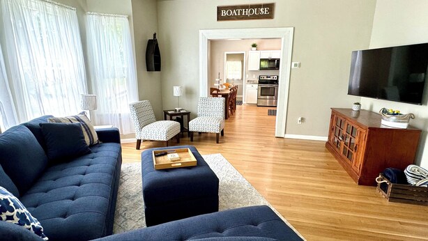 A living room with a sofa, ottoman, and chairs, a dining area with a table and chairs, and a kitchen with appliances. - Burnside Boathouse – Cozy 5 Bedroom Lake Retreat Steps from Marina & Local Eats (Burnside)