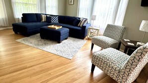 A living room with a sectional sofa, a patterned armchair, and a small ottoman. - Burnside Boathouse – Cozy 5 Bedroom Lake Retreat Steps from Marina & Local Eats (Burnside)