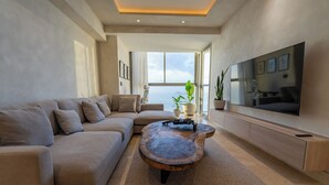 A modern living room with a sectional sofa, a wooden coffee table, a flat-screen TV, and a large window offering a view of the sky.
