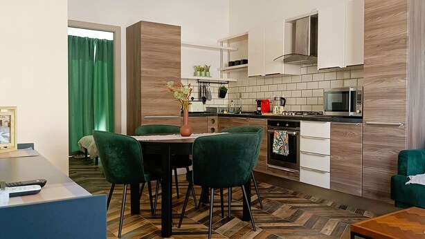 Une cuisine moderne avec un coin repas, des chaises vertes, une table et un canapé agrémenté d’un coussin à motifs floraux.