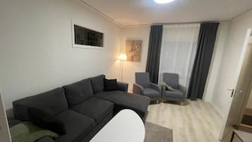 Un salon moderne comprenant un canapé d’angle, une table basse blanche, deux fauteuils, un lampadaire et une porte donnant sur une autre pièce.