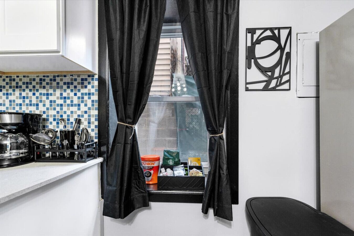 A compact kitchen with a window, black curtains, a coffee maker, and a tiled backsplash.