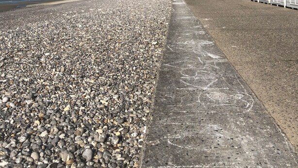 Une plage de galets avec une promenade en béton et, au fond, des cabanes de plage blanches.