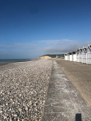 Une plage de galets avec une promenade en béton et, au fond, des cabanes de plage blanches.