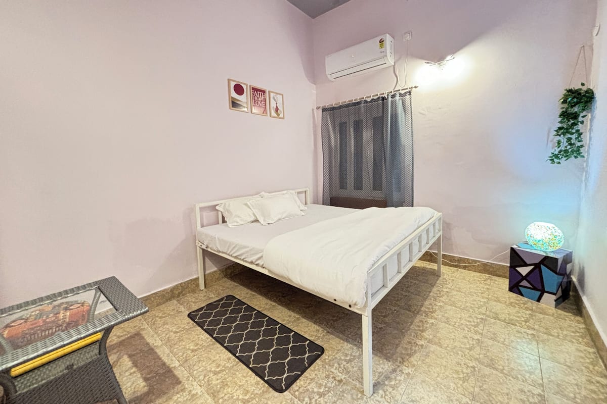 A bedroom with a bed, a small table, a mat, an air conditioner, and a wall-mounted light fixture.