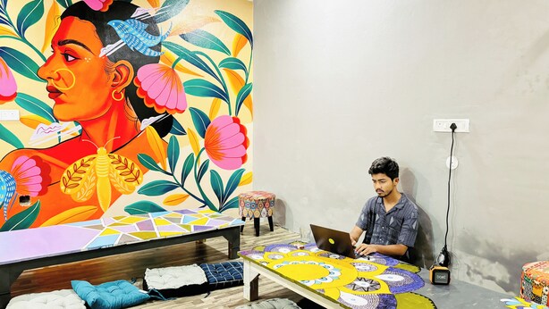 A room with a large colorful mural, a wooden bed, a small table with a laptop, and a power outlet on the wall. - Dormtales Hostel (Varanasi)