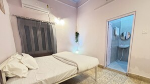 A bedroom with a bed, a small bedside table, a window with curtains, an air conditioning unit, and a bathroom visible through an open door. - Dormtales Hostel (Varanasi)