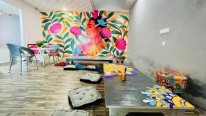 A room with a colorful mural, a wooden floor, a table with a patterned tablecloth, and a cushioned bench. - Dormtales Hostel (Varanasi)