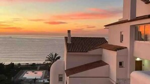 Un edificio costero con techo de tejas, piscina y vista al océano al atardecer.