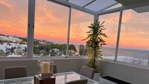 Un área de comedor con una mesa y sillas de vidrio, una planta en maceta y vistas al atardecer sobre el océano.