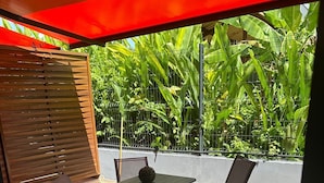 Um pátio externo com uma mesa e cadeiras, cercado por plantas verdes exuberantes e um portão de metal.