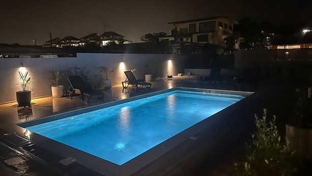 Piscina retangular com borda escura, cercada por espreguiçadeiras e plantas em vasos, iluminada à noite.