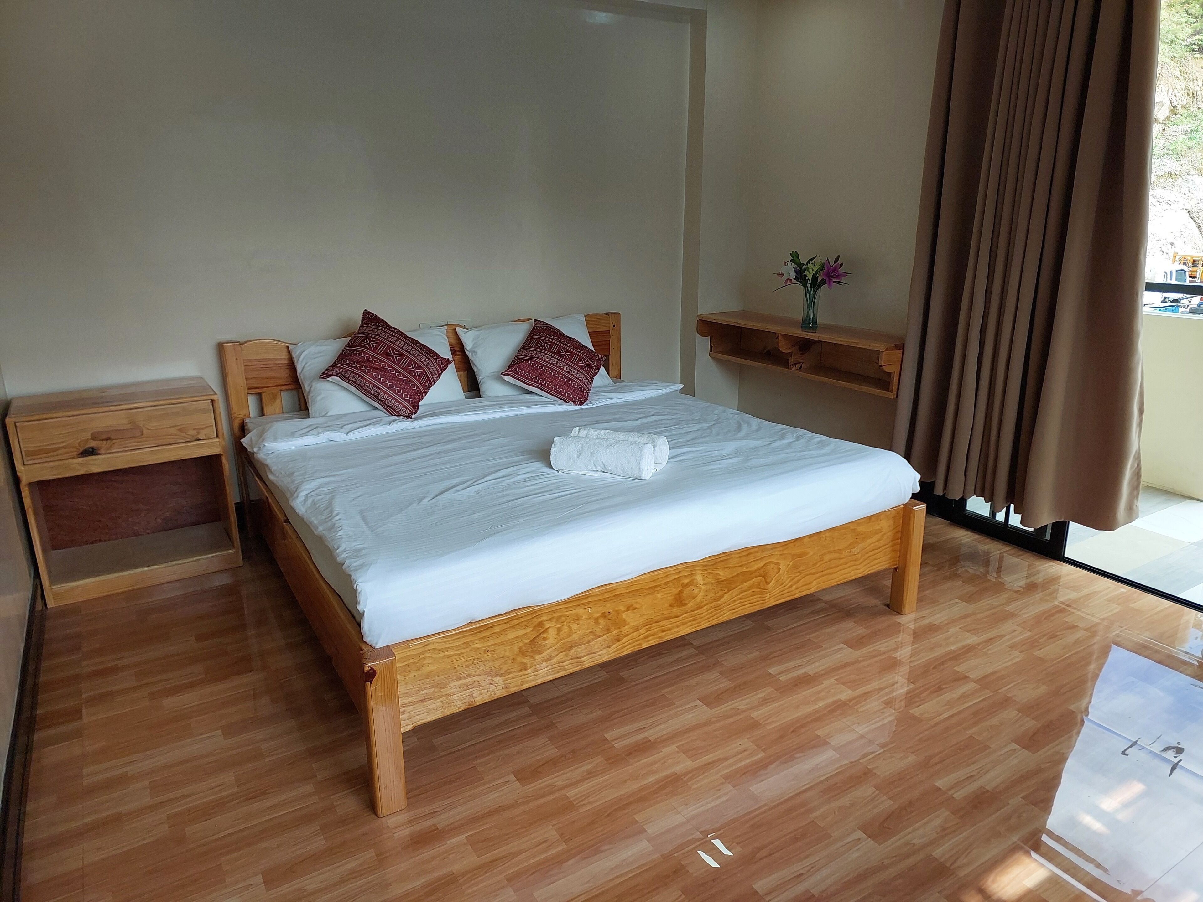Un lit bien fait, avec du linge de lit blanc et deux coussins décoratifs, une table de chevet en bois, une petite étagère avec un vase, et une vue sur l’extérieur par une fenêtre ornée de rideaux.