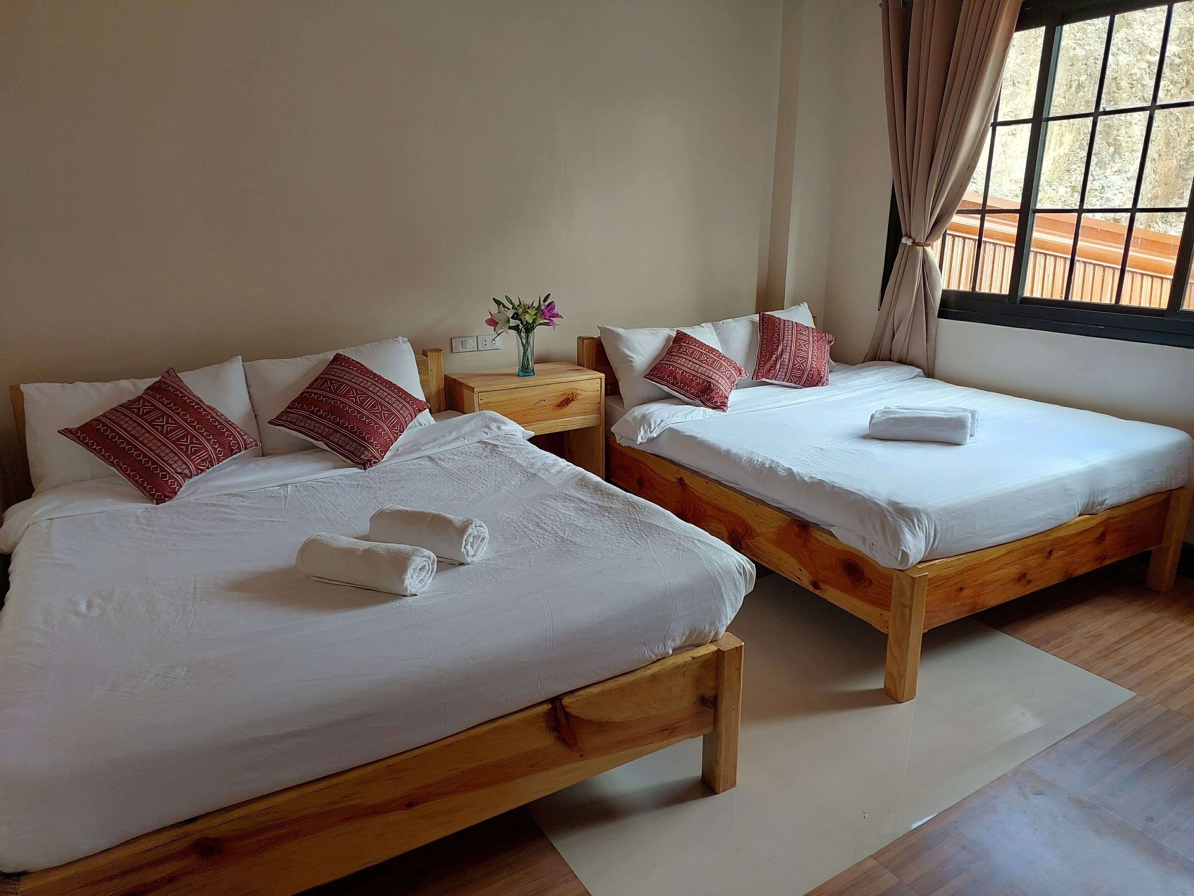 A hotel room with two wooden beds, white linens, red patterned pillows, a small wooden nightstand with a vase of flowers, and a window with curtains.