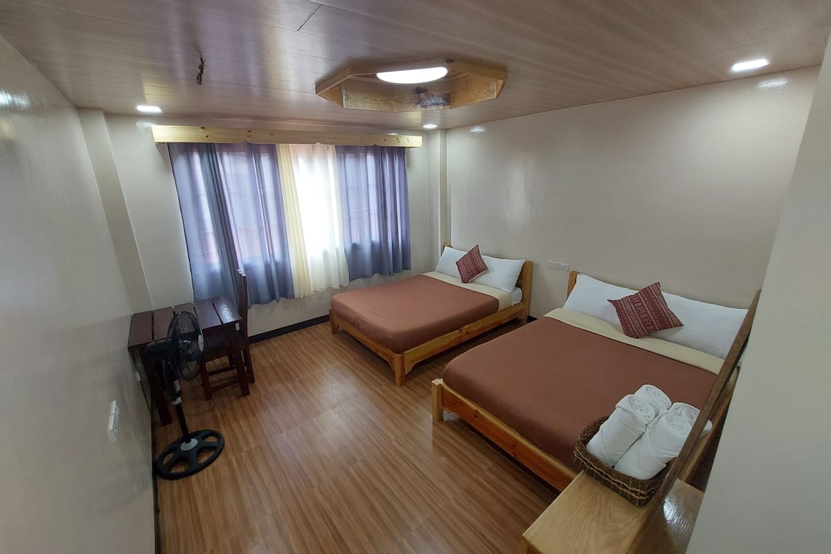 A hotel room with two single beds, a desk, a chair, and a fan.
