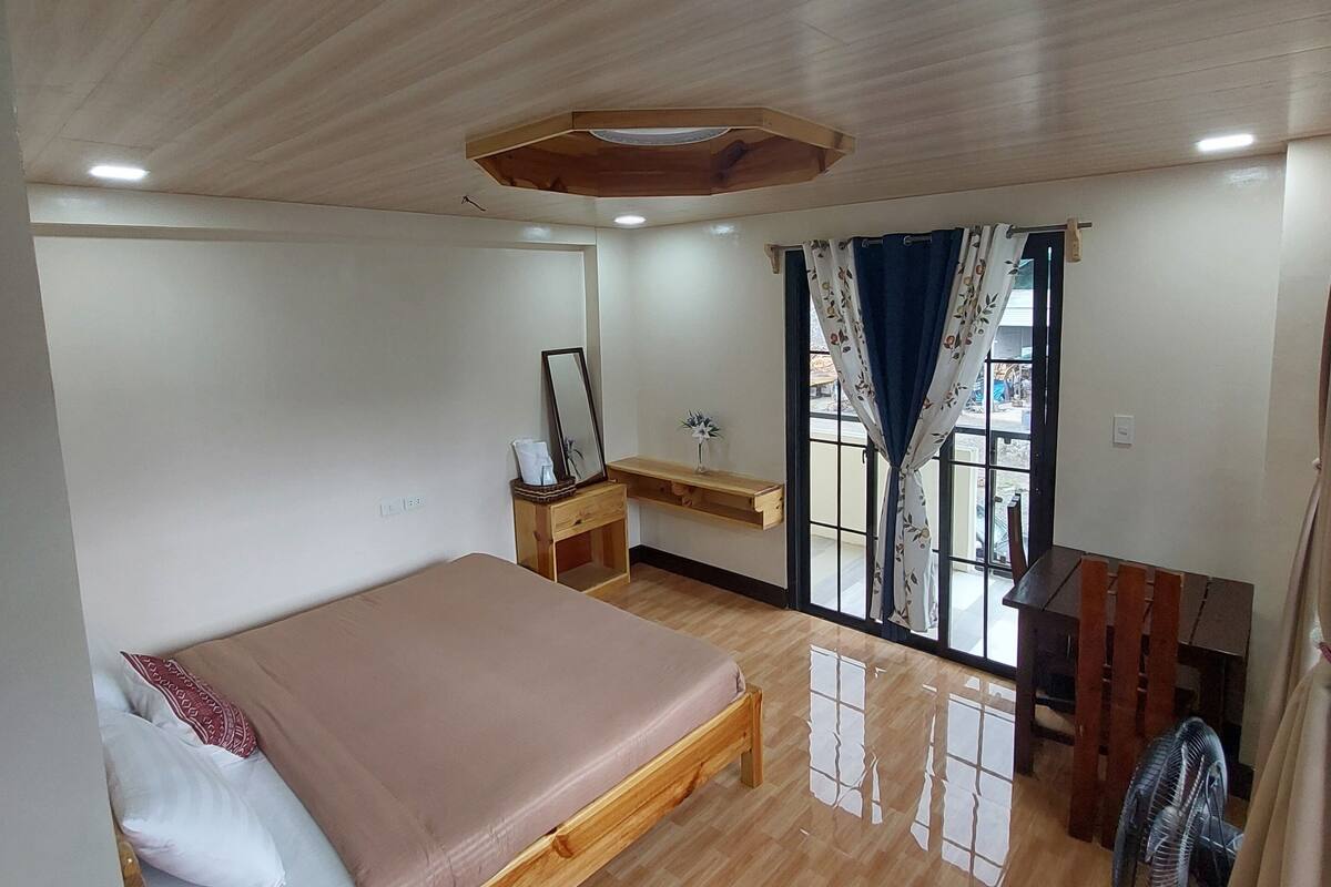 A bedroom with a wooden bed, a desk, and a chair. There is a window with curtains and a view outside.