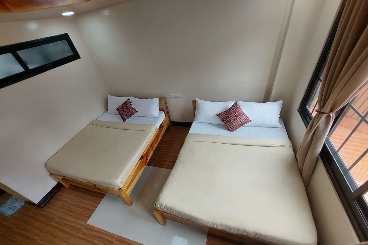 A compact room with two single beds, each with a wooden frame and white bedding. There is a small window with curtains, and the room has a tiled floor.