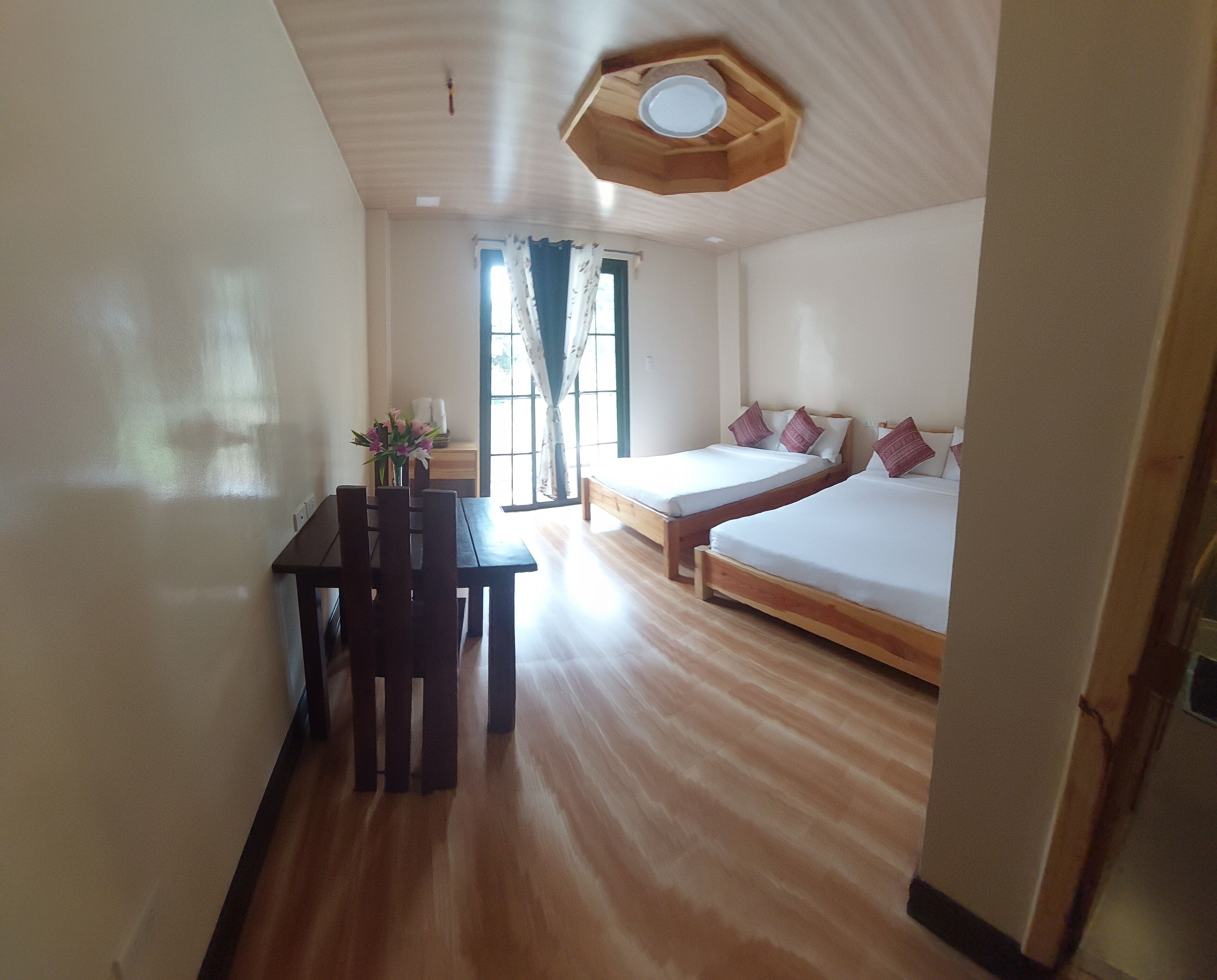 A hotel room with two single beds, a wooden table with chairs, a small desk, and a ceiling light fixture.
