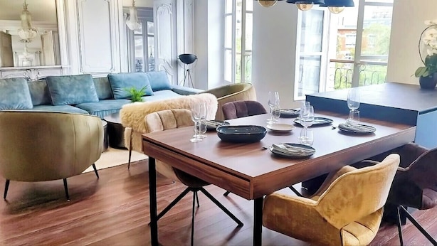 A dining area with a wooden table set for four, surrounded by chairs. A blue sofa and a small side table with a plant are visible in the background.