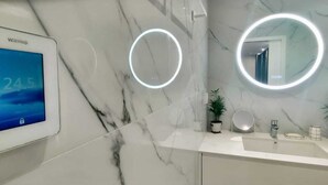 A bathroom with a digital thermostat displaying 24.5°C, a marble countertop, a sink, a mirror with a circular light, and potted plants.