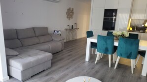 Una sala de estar moderna con un sofá gris, un comedor con sillas color turquesa y una cocina con armarios blancos.