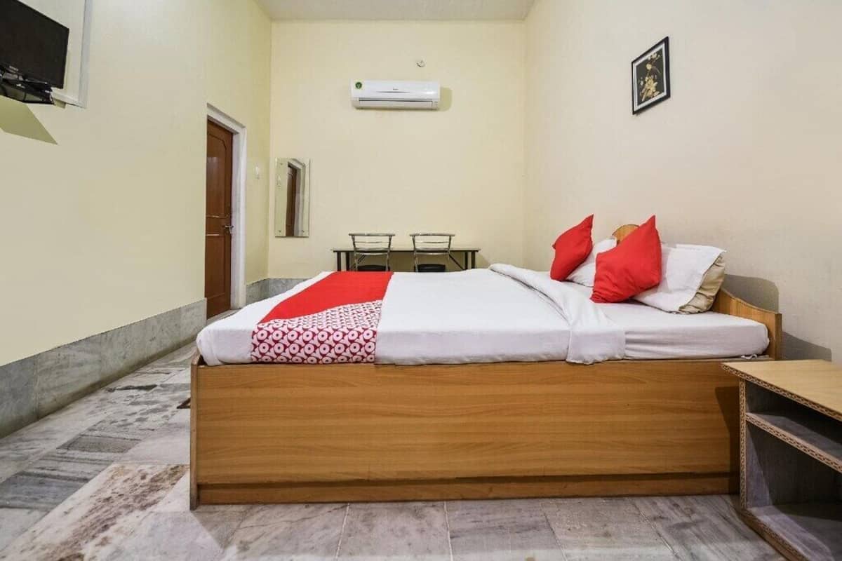 Un dormitorio con una cama de madera, una colcha de cuadros rojos y blancos, una pequeña mesa con sillas, un equipo de aire acondicionado y una puerta.