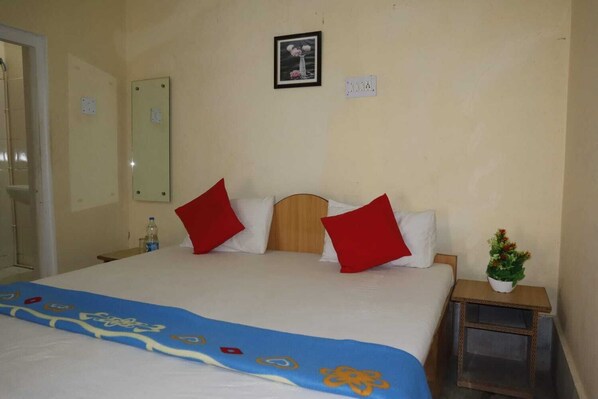 Una habitación de hotel con una cama, dos almohadas rojas, una colcha azul, una mesita con una planta y un cuadro en la pared.