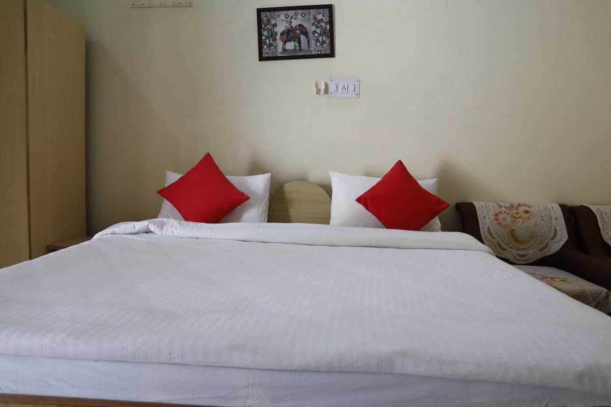 Una cama bien tendida con ropa blanca y dos almohadas rojas, un armario de madera a la izquierda, y un cuadro en la pared.