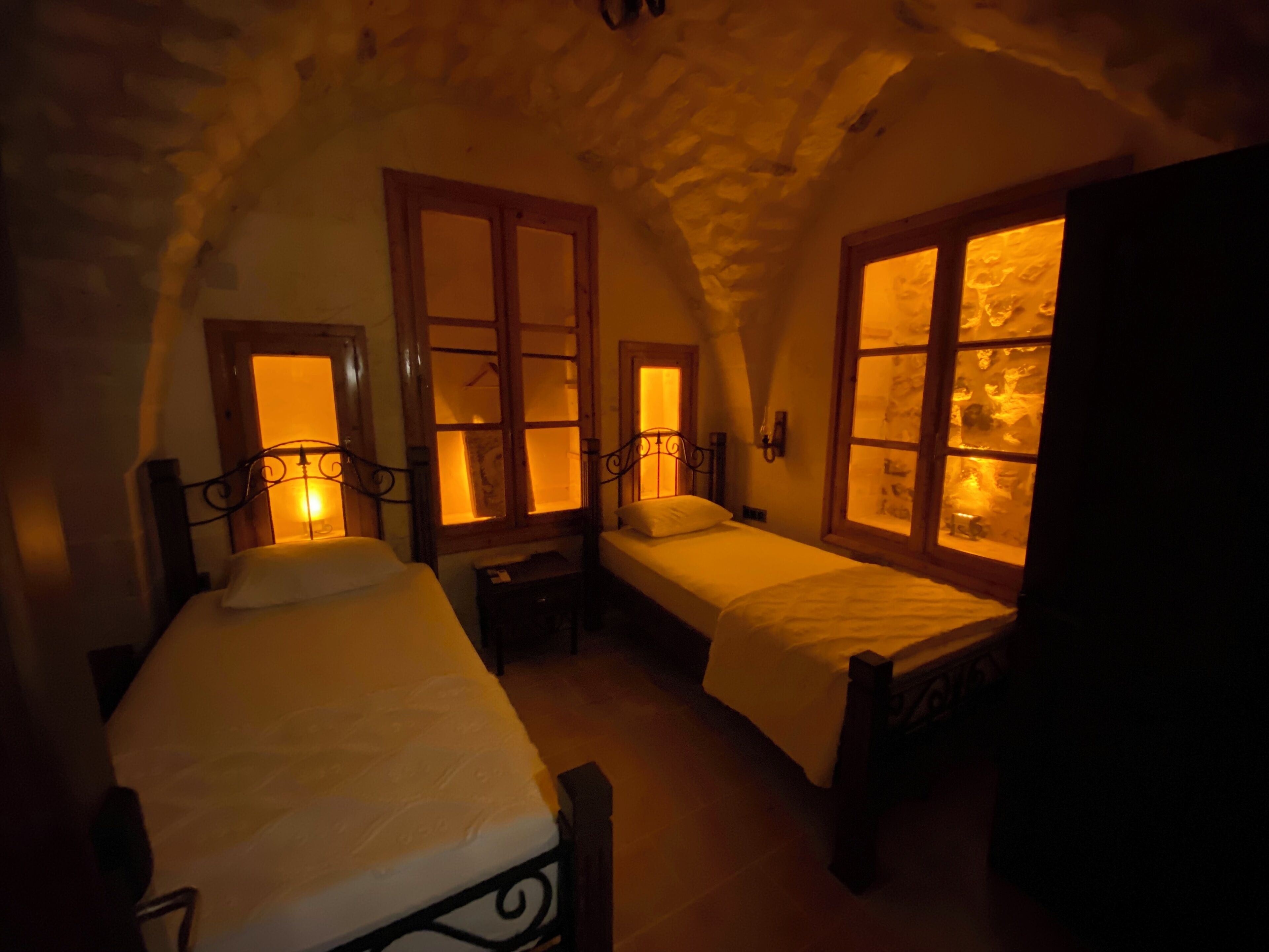 A cozy room with two beds, each with a wooden headboard and a window with a warm light. The room has a stone ceiling and a rustic, wooden floor.