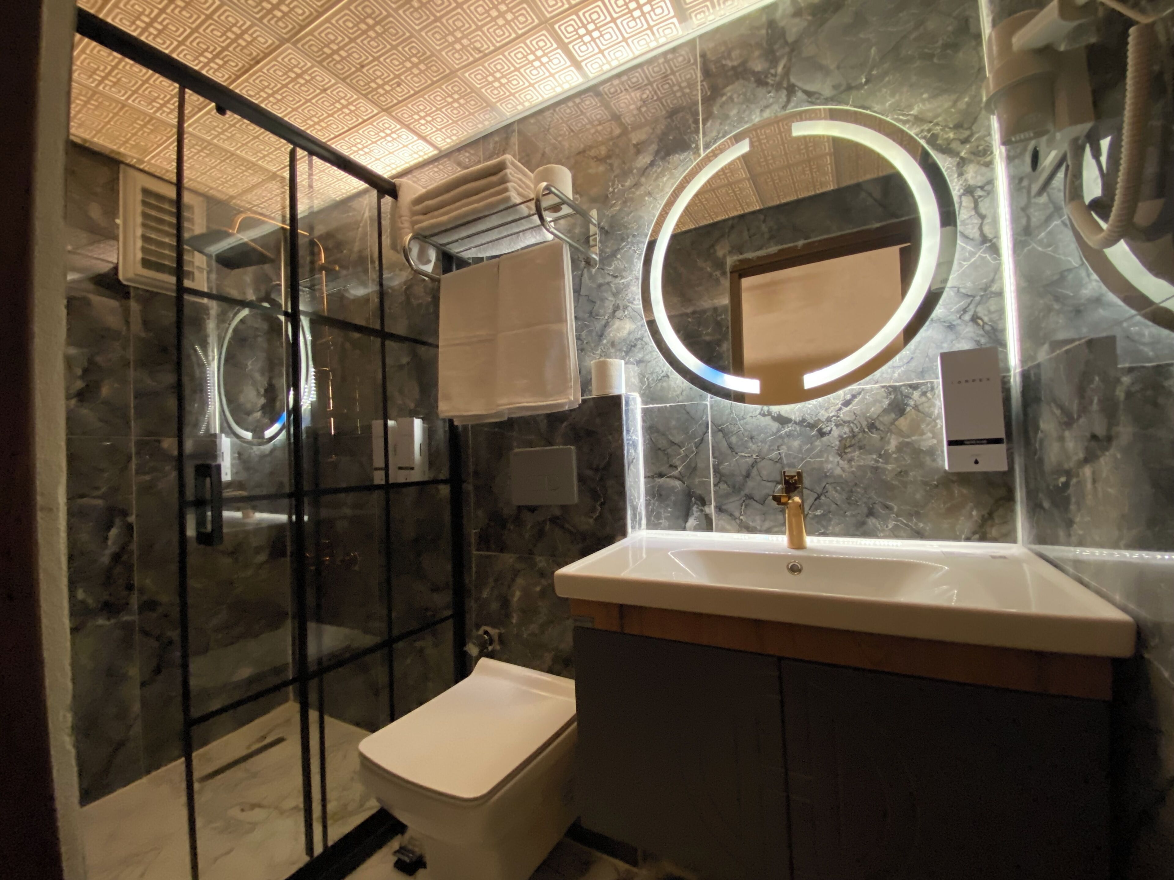 Un baño moderno con cabina de ducha de vidrio, inodoro blanco, tocador con encimera de mármol y un espejo grande con luz circular.