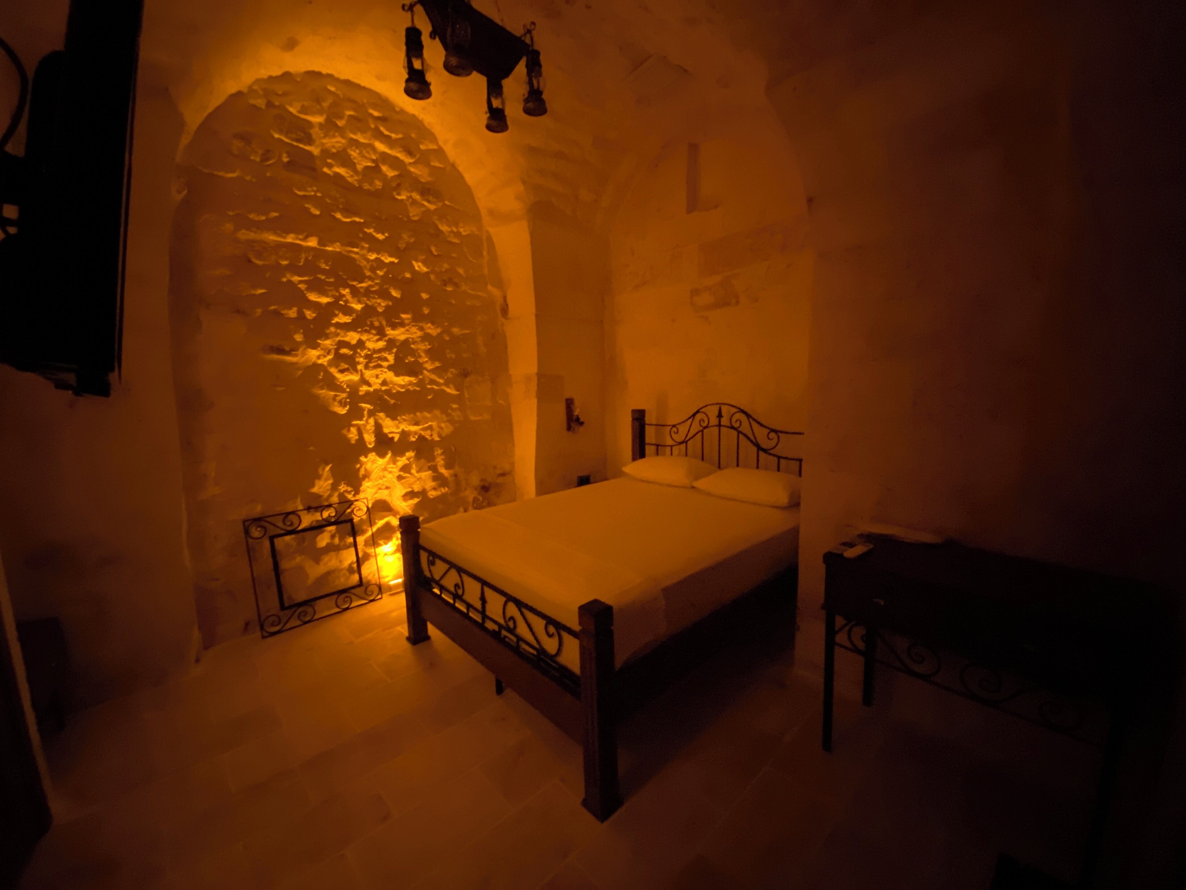 A dimly lit room with a stone wall, a bed with a metal frame, and a small table.
