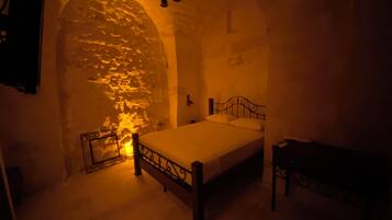 A dimly lit room with a stone wall, a bed with a metal frame, and a small table.