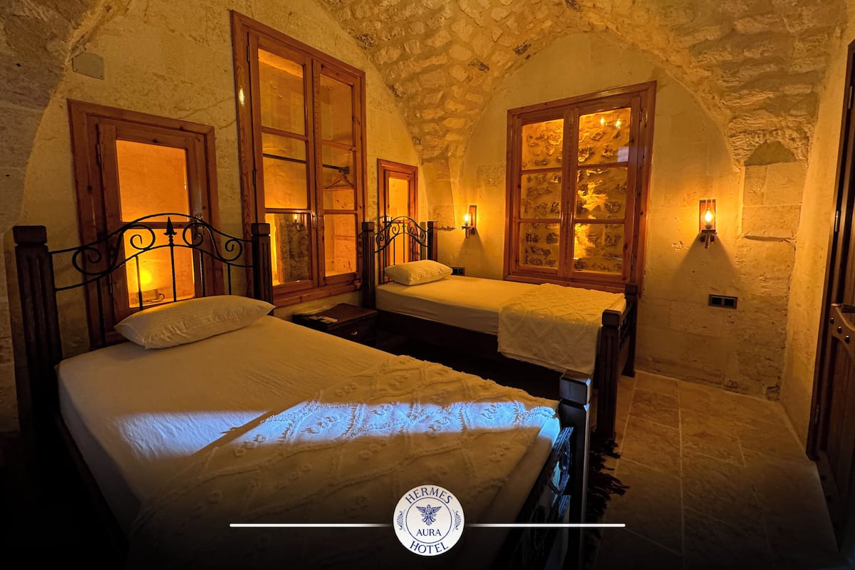 A room with two beds, stone walls, wooden beams, and wall-mounted lights.
