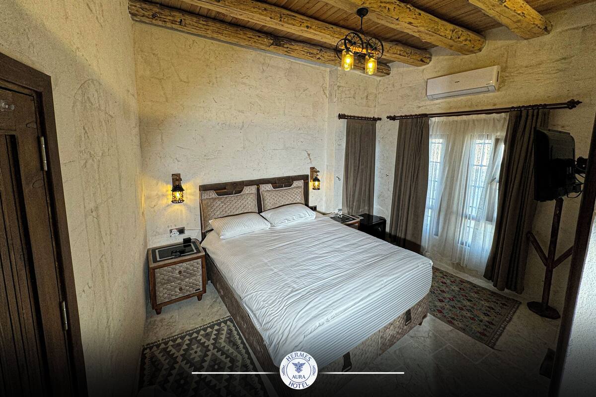 A bedroom with a wooden ceiling, a bed with a headboard, a nightstand, a wall-mounted TV, and a window with curtains.
