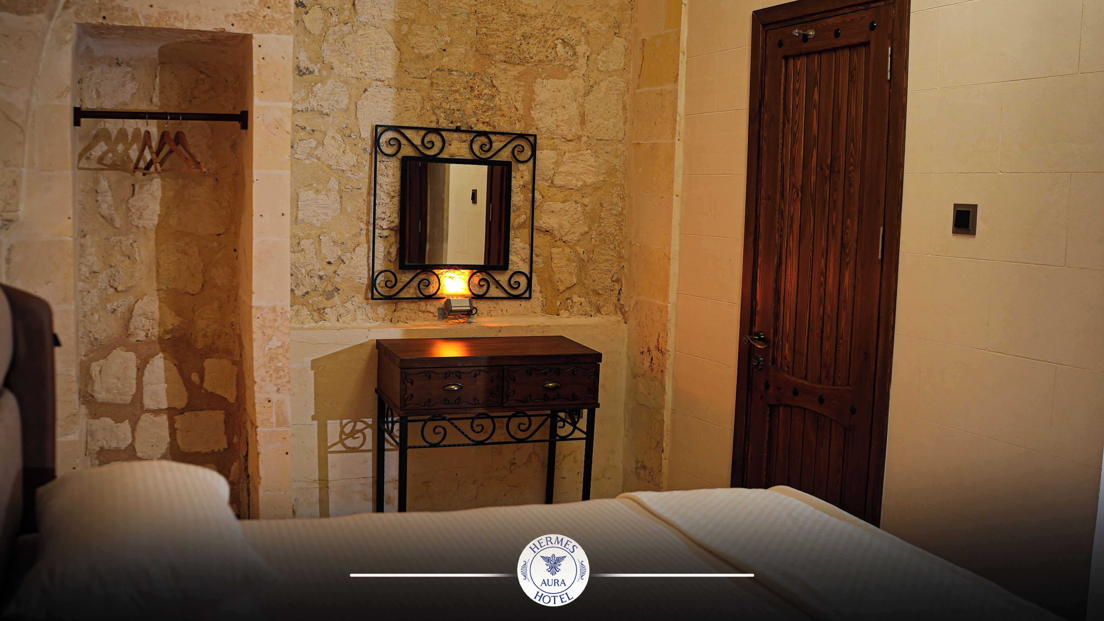 A hotel room with a bed, a wooden nightstand with a mirror, a wooden door, and a stone wall.