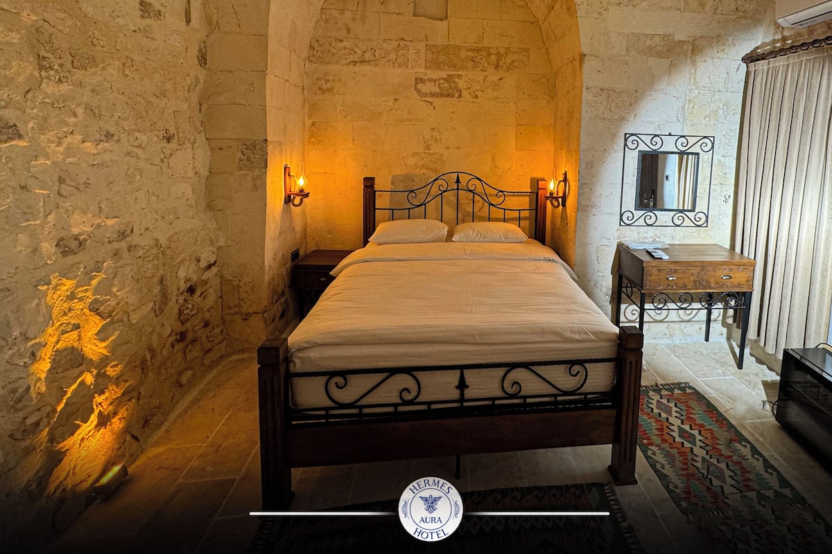 A bedroom with a stone wall, a wooden bed, a nightstand, a mirror, and a small table.